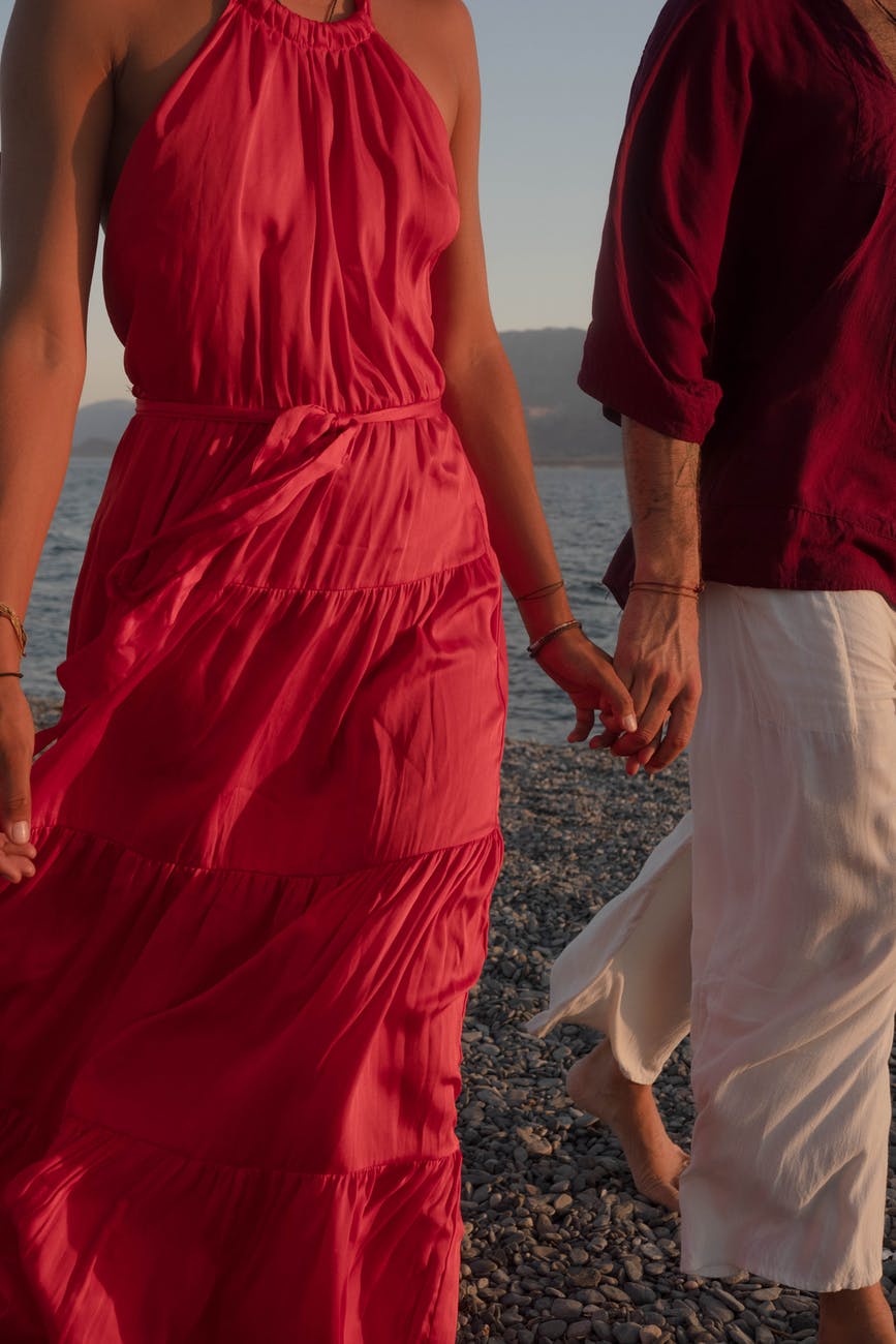 man and woman in red dress walking hand in hand in the beach