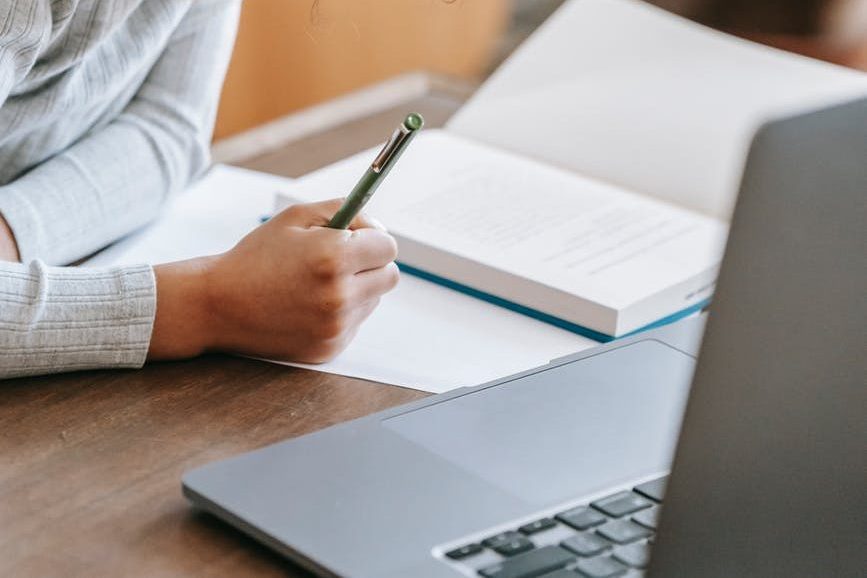 focused black schoolkid taking notes during online lesson via laptop; writing books: Laura