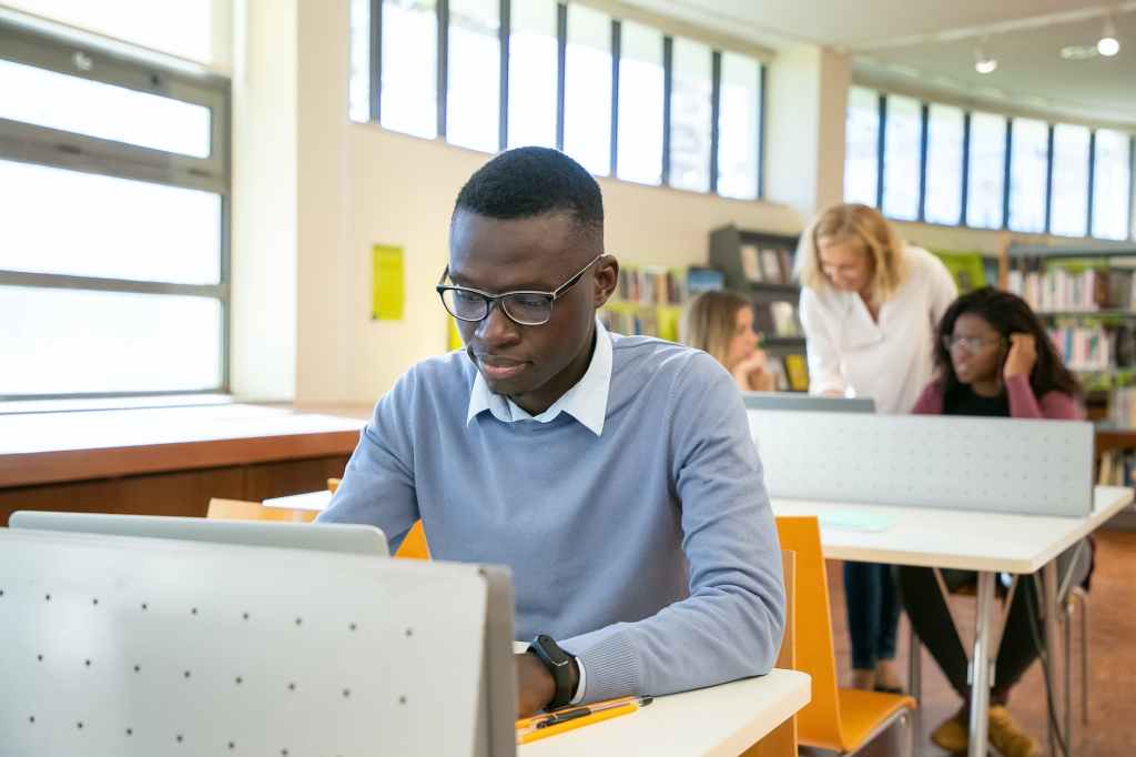 smart young ethnic guy browsing laptop during lesson in university; digital library